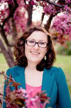 An image of a woman with short, curly brown hair, wearing a teal blazer with a pink shirt underneath. She has light skin and wears glasses. She is smiling. She is outdoors and standing between tree branches that have pink flowers.